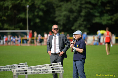 Zwei Personen halten neben aufgereihten Hürden eine Ansprache ins Mikrofon beim Walfried-Heinz-Sportfest der TG Konz im Saar-Mosel-Stadion in Konz.