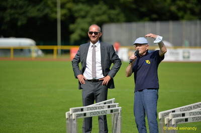 Eine Person mit Mikrofon und eine Person im Anzug stehen neben aufgereihten Hürden beim Walfried-Heinz-Sportfest der TG Konz im Saar-Mosel-Stadion in Konz.