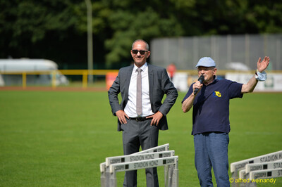 Zwei Personen begrüßen mit Mikrofon die Teilnehmenden neben aufgereihten Hürden beim Walfried-Heinz-Sportfest der TG Konz im Saar-Mosel-Stadion in Konz.