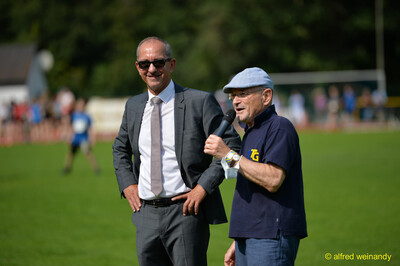 Eine Person mit Mikrofon und eine Person im Anzug stehen lachend auf dem Rasen beim Walfried-Heinz-Sportfest der TG Konz im Saar-Mosel-Stadion in Konz.
