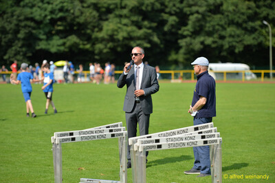 Eine Person im Anzug spricht ins Mikrofon neben aufgereihten Hürden beim Walfried-Heinz-Sportfest der TG Konz im Saar-Mosel-Stadion in Konz.