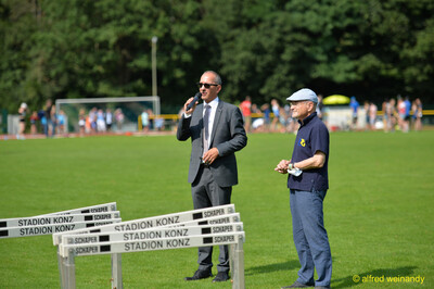 Eine Person im Anzug hält eine Ansprache ins Mikrofon neben aufgereihten Hürden beim Walfried-Heinz-Sportfest der TG Konz im Saar-Mosel-Stadion in Konz.