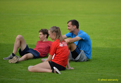 Drei junge Menschen sitzen auf dem Rasen und verfolgen die Wettkämpfe beim Walfried-Heinz-Sportfest der TG Konz im Saar-Mosel-Stadion in Konz.