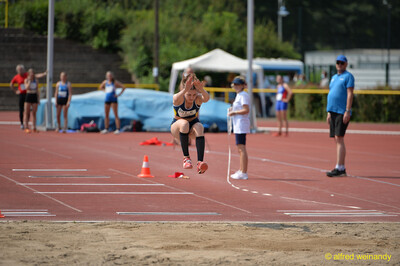 Eine Person springt in der Flugphase mit angezogenen Beinen Richtung Sandgrube beim Walfried-Heinz-Sportfest der TG Konz im Saar-Mosel-Stadion in Konz.
