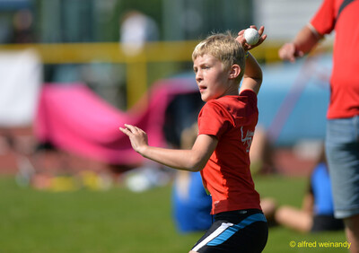 Ein blondes Kind im roten Trikot holt mit einem kleinen Schlagball zum Wurf aus bei der Rheinland-Meisterschaft im Mehrkampf der TG Konz in Konz.