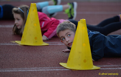 Mehrere Kinder liegen zum Aufwärmen hinter gelben Markierungskegeln auf der Laufbahn beim Jugendsportfest der TG Konz in Konz.