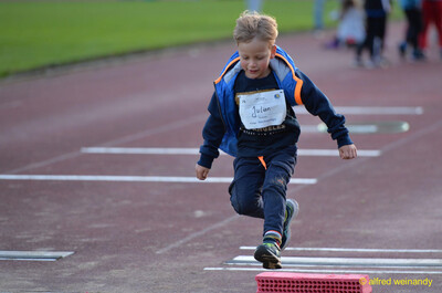 Ein Kind springt bei einer Sprungübung über einen niedrigen roten Stein auf der Laufbahn beim Jugendsportfest der TG Konz in Konz.