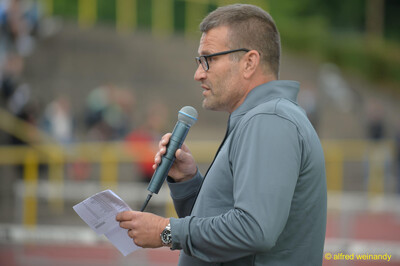Eine Person an der Stadionansage verkündet mit Mikrofon und Ergebnisblatt die Resultate bei der Rheinland-Pfalz-Meisterschaft in Konz.