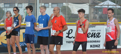 Mehrere Aktive stehen mit Startnummern nebeneinander an der Bande und warten auf ihren Wettkampf beim Walfried-Heinz-Sportfest der TG Konz im Saar-Mosel-Stadion in Konz.