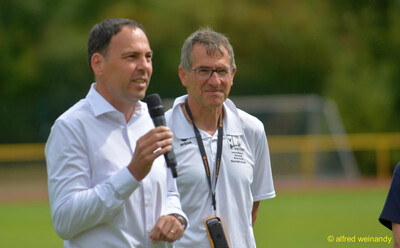 Eine sprechende Person und eine Person im Kampfgericht stehen auf dem Sportplatz beim Walfried-Heinz-Sportfest der TG Konz im Saar-Mosel-Stadion in Konz.