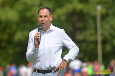 Eine Person im weißen Hemd hält eine Ansprache mit dem Mikrofon beim Walfried-Heinz-Sportfest der TG Konz im Saar-Mosel-Stadion in Konz.