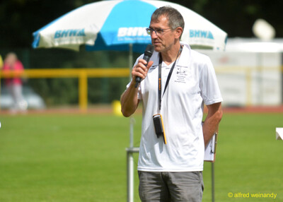 Eine Person im Kampfgericht spricht ins Mikrofon vor einem Sonnenschirm beim Walfried-Heinz-Sportfest der TG Konz im Saar-Mosel-Stadion in Konz.