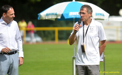 Zwei Personen stehen vor einem Sonnenschirm, eine spricht ins Mikrofon, beim Walfried-Heinz-Sportfest der TG Konz im Saar-Mosel-Stadion in Konz.