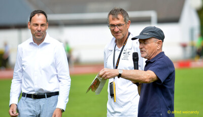 Drei Personen stehen im Gespräch auf dem Rasen, eine hält Mikrofon und Programmblatt, beim Walfried-Heinz-Sportfest der TG Konz im Saar-Mosel-Stadion in Konz.