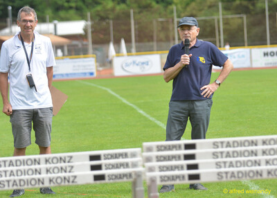 Zwei Vereinsverantwortliche stehen mit Mikrofon auf dem Rasen vor gestapelten Hürden beim Walfried-Heinz-Sportfest der TG Konz im Saar-Mosel-Stadion in Konz.