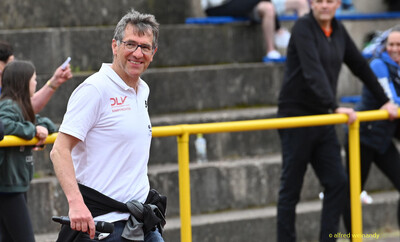 Eine lächelnde Person im Kampfgericht mit Mikrofon geht vor der Tribüne entlang beim Walfried-Heinz-Sportfest der TG Konz im Saar-Mosel-Stadion in Konz.