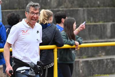 Eine lachende Person im Kampfgericht mit Mikrofon steht am Bahngeländer beim Walfried-Heinz-Sportfest der TG Konz im Saar-Mosel-Stadion in Konz.