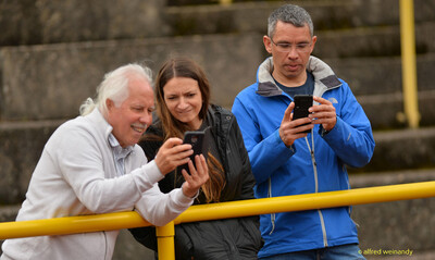 Drei Personen betrachten lächelnd ihre Smartphones an der Tribünenabsperrung beim Walfried-Heinz-Sportfest der TG Konz im Saar-Mosel-Stadion in Konz