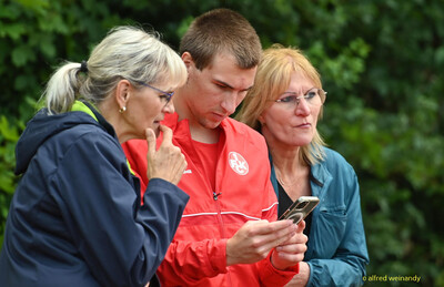 Drei Personen schauen zusammen auf ein Smartphone am Rand der Sportanlage bei der Rheinland-Meisterschaft im Mehrkampf der TG Konz in Konz.