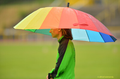 Eine Person steht mit einem bunten Regenschirm am Rand der Anlage bei der Rheinland-Meisterschaft im Mehrkampf der TG Konz in Konz.