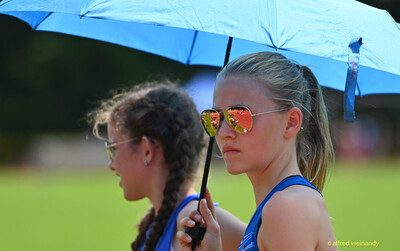 Zwei Jugendliche warten unter einem blauen Regenschirm im Schatten bei der Rheinland-Meisterschaft im Mehrkampf der TG Konz in Konz.