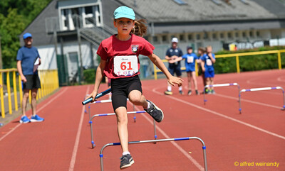 Ein Kind überquert mit dem Staffelstab in der Hand eine Hürde auf der Laufbahn beim Kinderleichtathletik-Sportfest der TG Konz im Saar-Mosel-Stadion in Konz