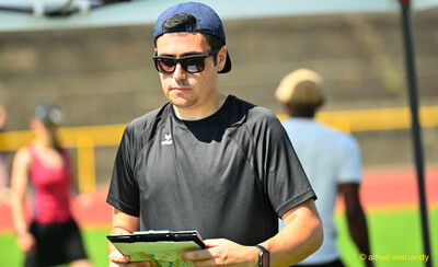 Eine Person mit Kappe und Sonnenbrille prüft eine Ergebnisliste auf einem Klemmbrett beim Kinderleichtathletik-Sportfest der TG Konz im Saar-Mosel-Stadion in Konz
