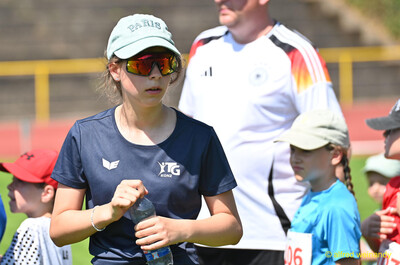 Eine Person mit Sonnenbrille und Wasserflasche steht zwischen Kindern beim Kinderleichtathletik-Sportfest der TG Konz im Saar-Mosel-Stadion in Konz